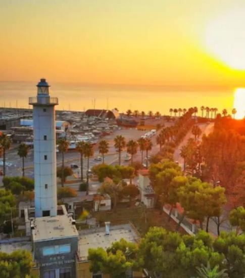 Tramonto sul faro e il porto, circondati da palme e mare calmo.